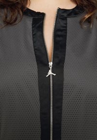 Gros plan d'une veste noire zippée en tissu perforé avec une tirette de fermeture éclair en forme de logo de joueur de basket.