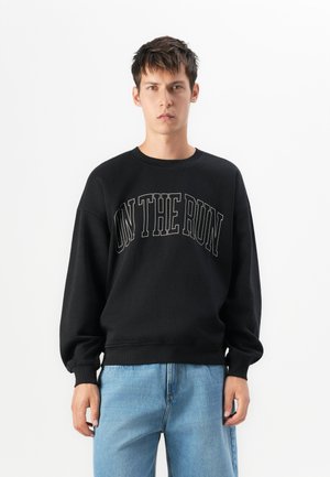 Jonge man die tegen een witte achtergrond staat, gekleed in een zwart sweatshirt met de tekst "ON THE RUN" en lichtblauwe spijkerbroek.