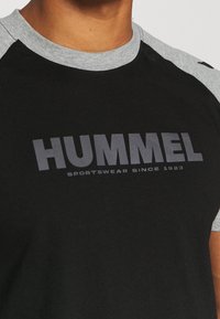 Sort T-shirt med korte ærmer og grå raglan ærmer. Har "HUMMEL" logoet og teksten "SPORTSWEAR SINCE 1923" i mørkegrå skrifttype.