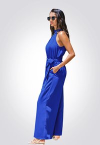 Mono azul sin mangas con cinturón atado, diseño de pierna ancha y dos bolsillos laterales. La modelo lo combina con sandalias blancas de tiras y gafas de sol.