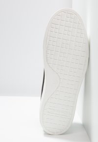 Semelle de basket blanche avec un motif de bande de roulement en forme de grille reposant contre un coin de mur et de sol blanc.