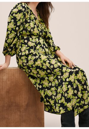 Robe fleurie en tissu noir, ornée de fleurs vertes et crème, avec un décolleté en V et des manches trois-quarts, tombant jusqu'au mollet.