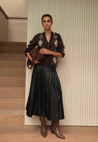 Brown argyle-patterned cardigan over a pleated black skirt, paired with brown heeled boots. A brown bag is held in the left hand.