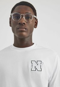Biała koszula z długim rękawem i okrągłym dekoltem, z czarnym haftowanym logo "N". Przezroczyste okrągłe okulary dopełniają całość.