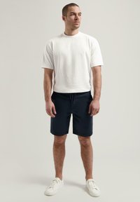 Weißes T-Shirt mit kurzen Ärmeln, kombiniert mit blauen Shorts mit Kordelzug und abgerundet mit weißen Sneakers.