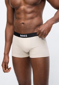 Μυώδης άνδρας που προσαρμόζει μπεζ μποξεράκια με μαύρη ζώνη μέσης με την ένδειξη "BOSS" μπροστά σε απλό φωτεινό φόντο.