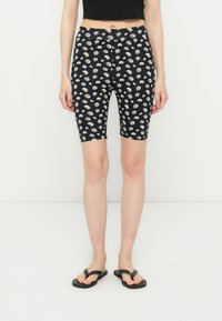 Γυναίκα που φορά μαύρα σορτς μέχρι τα μέσα του μηρού με λευκή στάμπα με μαργαρίτες, μαύρο κοντό τοπ και μαύρες σαγιονάρες, στέκεται μπροστά σε λευκό φόντο.