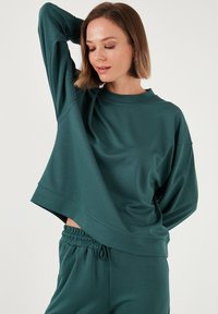 Felpa oversize color teal realizzata in tessuto morbido e liscio. Presenta un collo alto e spalle scese, da abbinare a pantaloni jogger coordinati.