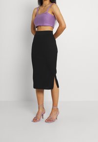 Schwarzer gerippter Midi-Rock mit Seitenschlitz, kombiniert mit einem lila gestrickten Crop-Top und lila Riemchen-High-Heels. Glatte Textur und figurbetonte Silhouette.