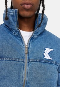 Blue denim jacket with high collar, front zipper, and two chest pockets. Features white embroidered logo on the left side.