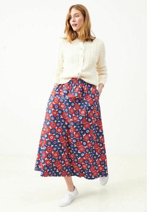 Nainen, jolla on valkoinen neulecardigan, tummansininen hame, jossa punavalkoinen kukkakuvio, ja valkoiset tennarit, seisoo kädet taskuissa.