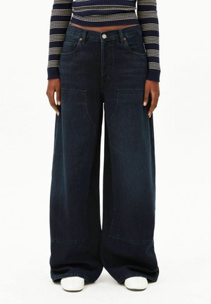 Vrouw draagt donkere, wijd uitlopende jeans met grote voorzakken, een gestreepte cropped trui en witte schoenen, staand tegen een witte achtergrond.
