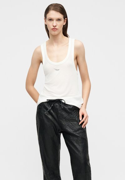 Γυναίκα που φοράει λευκό αμάνικο τοπ με μικρό λογότυπο και μαύρο υφασμάτινο παντελόνι με κορδόνι, στέκεται με τα χέρια στις τσέπες.