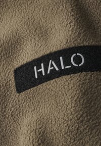 Lysebrunt fleece-stof med en tekstureret overflade, der har et sort rektangulært patch mærket "HALO" med hvide bogstaver.