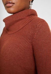 Gros plan sur une personne portant un pull turtleneck en tricot épais de couleur rouille et des boucles d'oreilles en argent, montrant les détails de la texture du tissu.