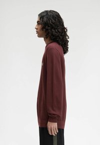 Giovane con lunghi capelli ricci, che indossa un maglione bordeaux e pantaloni neri, ritratto di profilo su uno sfondo bianco uniforme.