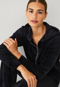 Zwarte velours trainingspak met een rits hoodie, met een gladde textuur en subtiele glans. De handen van het model zijn ontspannen gekruist.