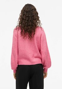 Femme avec de longs cheveux bouclés portant un pull rose à manches longues et un pantalon noir, se tenant de dos contre un fond blanc.