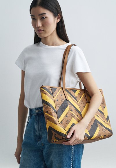 Naine kannab valget T-särgiga ja siniste teksadega suurt pruunikaskollase ja musta mustriga kotti, millel on nahast sangad õlal.