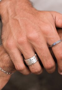 Silver rings on two hands: one is wide with an engraved floral pattern, the other is slender with a blue stone, both textured.