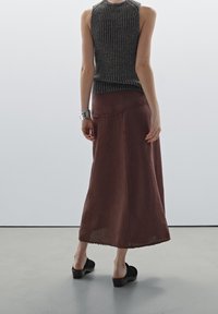 Nainen, joka on pukeutunut hihattomaan tummanraitoiseen paitaankin, pitkään ruskeaan hameeseen, jossa on raaka helma, metalliseen rannerenkaaseen ja mustiin korkokenkiin, seisoo harmaalla lattialla.