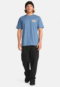 Tricou albastru din bumbac cu mâneci scurte, având un mic grafic cu val pe buzunarul de la piept. Asortat cu pantaloni largi negri și adidași negri.