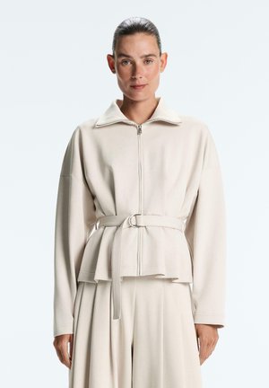 Nő világos bézs színű, cipzáras, övvel ellátott dzsekit és hozzá illő bő szárú nadrágot visel, egyszerű, világos háttér előtt áll.