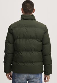 Dark green puffer jacket with a high collar, quilted design, and an elasticized cuff detail; paired with light blue denim jeans.