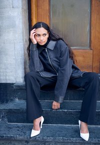 Kobieta z długimi ciemnymi włosami w czarnej kurtce, czarnych spodniach i białych szpilkach siedzi na kamiennych schodach, opierając głowę na dłoni.