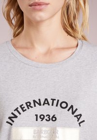 Zblízka na osobu, která má na sobě šedou košili s černým potiskem "INTERNATIONAL 1936" a reflexní štítek "BARBOUR INTERNATIONAL".