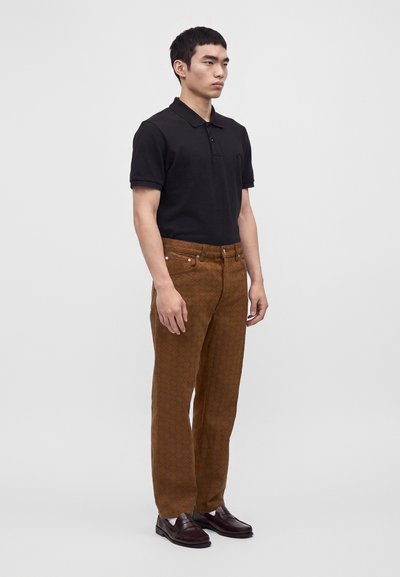 Jeune homme debout, portant un polo noir à manches courtes, un pantalon marron à motifs, des chaussettes blanches et des mocassins marron foncé, sur un fond uni.