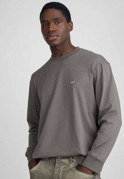 Pelēkais garo piedurknju krekls ar noapaļotu kakla izgriezumu un mazu logotipu uz krūtīm, izgatavots no mīkstas, teksturētas auduma.