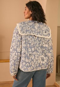 Giacca trapuntata con un motivo floreale blu e contorni bianchi. Presenta un ampio colletto con bordi arricciati. Indossata sopra jeans larghi.