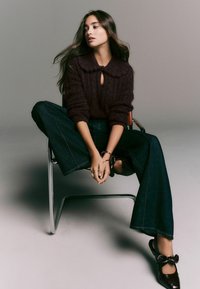 Pulover tricotat de culoare vișinie cu guler în formă de cheie, asociat cu blugi din denim închisi la culoare, cu pantaloni largi și pantofi maro cu accente sub formă de fundițe.