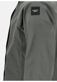 Giacca leggera di colore grigio scuro con una texture liscia. Presenta un'etichetta nera quadrata con la scritta "SWEDEMOUNT" e il logo di una montagna sulla manica.