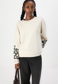 Beige genser med svart virvlende broderi på ermene, med rund hals og ribbestrikkede ermer, sammen med mørke jeans.