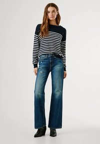 Temno modro-beli pulover z dolgimi rokavi z navpičnimi črtami, kombiniran z modrimi kavbojkami z visokim pasom in širokimi nogami, ki imajo obrabljene kolena, ter črnimi gležnjarji.