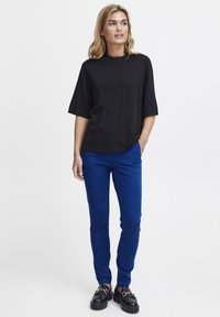 Schwarzes kurzärmliges Oberteil kombiniert mit blauen schmal geschnittenen Hosen und schwarzen klobigen Schuhen. Das Outfit zeichnet sich durch eine lockere Passform und glatte Textur aus.