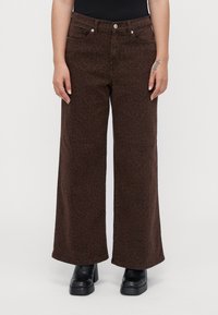 Persoon in een paarse wijde broek met een subtiele luipaardprint, zwarte platformboots en een zwart topje, staande tegen een effen achtergrond.