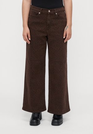 Persona vistiendo pantalones de pierna ancha marrones con un sutil estampado de leopardo, botas de plataforma negras y una parte superior negra, de pie contra un fondo sencillo.