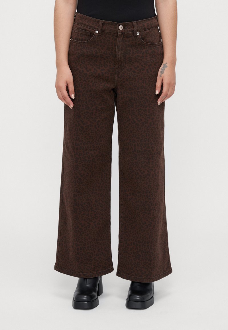 Persoon in een paarse wijde broek met een subtiele luipaardprint, zwarte platformboots en een zwart topje, staande tegen een effen achtergrond.