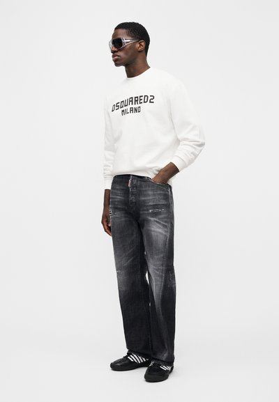 White sweatshirt with black logo, paired with dark gray distressed jeans. Model wears dark sunglasses and black sneakers with white stripes.