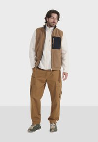 Chaleco de forro polar marrón sobre una chaqueta blanca con cremallera, combinado con pantalones cargo marrones. Tejido texturizado y detalle de bolsillo en negro. Zapatillas verdes.
