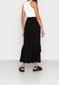 Jupe maxi noire en couches avec un tissu texturé, une taille élastique et un design fluide. Portée avec un débardeur blanc et des sandales noires épaisses.