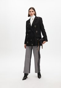 Blazer nero doppiopetto con decorazioni a forma di cuore, abbinato a pantaloni grigi a gamba larga. La modella tiene una piccola borsa nera.
