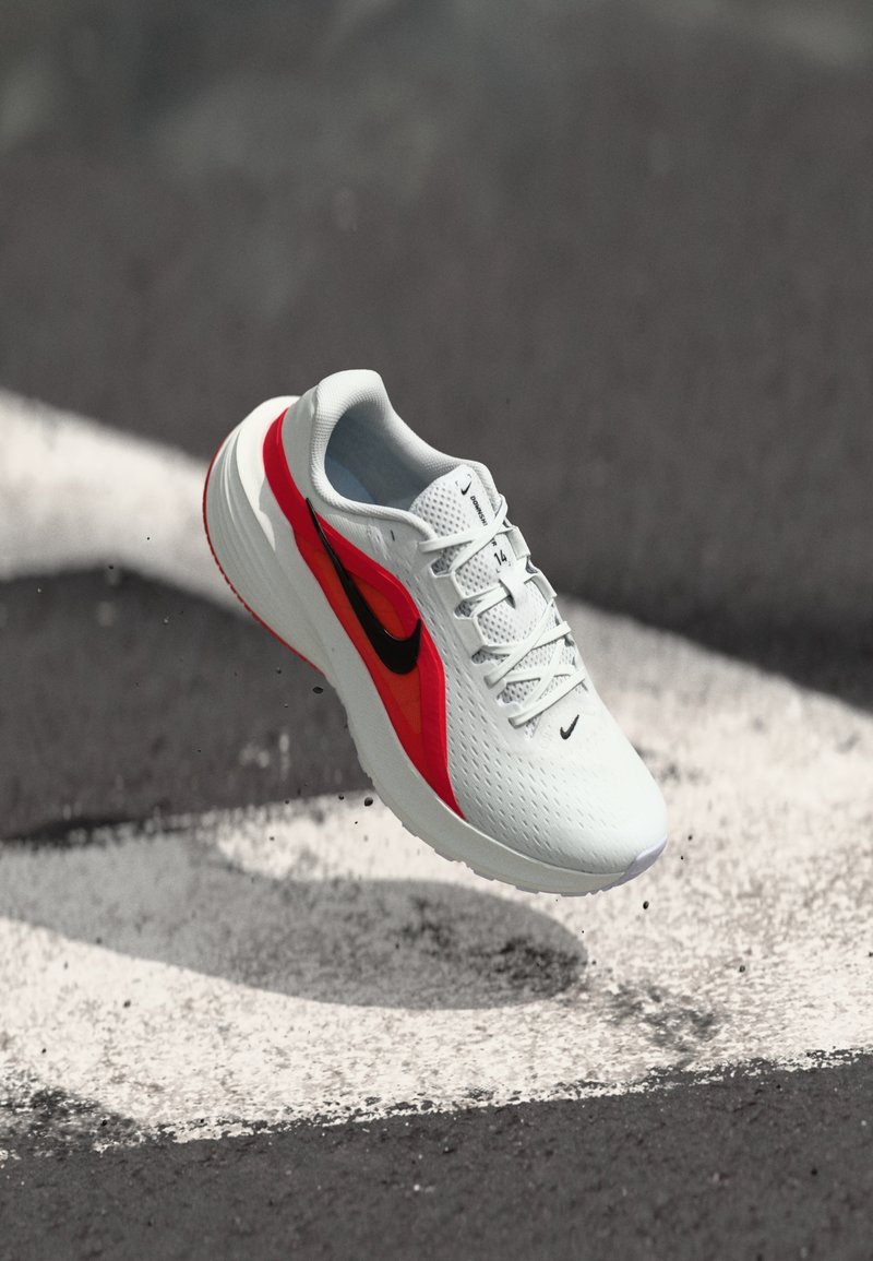 Chaussure de course blanche avec des touches rouges et noires, flottant au-dessus d'un bitume avec des marquages routiers blancs et des petits graviers dispersés.