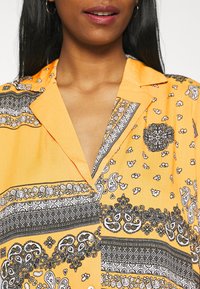 Camicia gialla con motivi paisley e floreali neri, maniche corte, colletto e chiusura a bottoni. Tessuto liscio e leggero con disegni intricati.