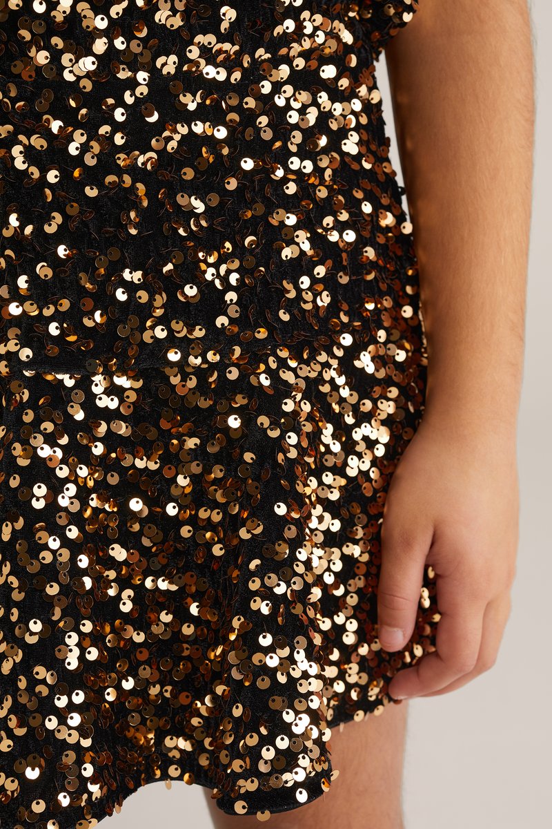 Schwarzes Kleid mit goldenen Pailletten, kurzem, gerafftem Saum und strukturierter Oberfläche. Eine Hand hält den Stoff und hebt seinen Glanz hervor.