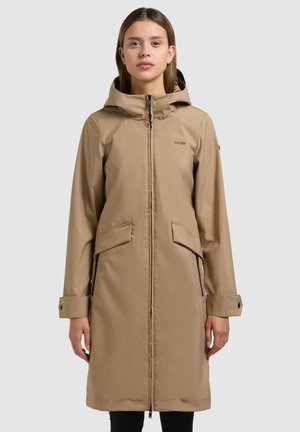 Woman wearing a knee-length beige hooded coat with front zipper and flap pockets, standing against a plain light background.