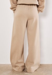 Pantaloni della tuta lunghi beige con una vestibilità ampia, vita elastica e texture morbida, che si allargano a forma di campana sul fondo.